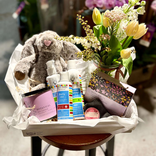 A curated gift basket or hamper containing a plush bunny toy, shower steamers, A candle, baby products and  a onesie with sock. Included is a posy of flowers in a bottle jar all arranged in a white paper-lined tray.