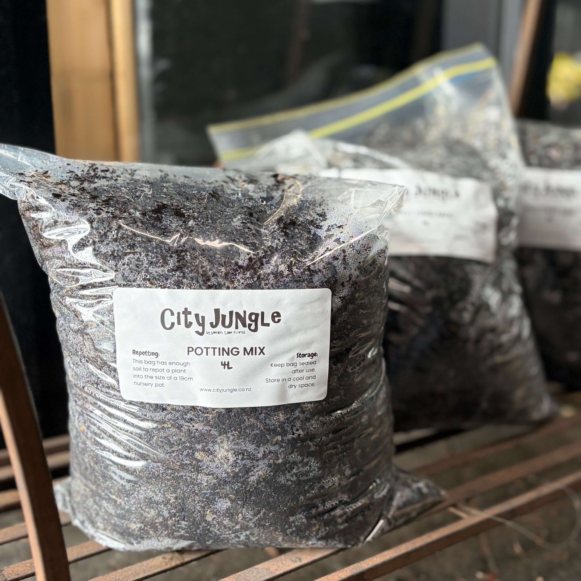 A clear plastic bag filled with dark potting mix sits on a wooden shelf. The front bag has a white label reading 'City Jungle' and 'POTTING MIX 4L'.